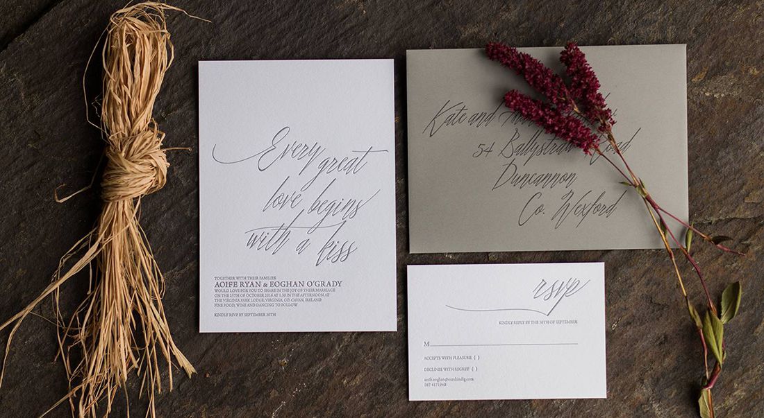 Wedding invitation and RSVP printed with grey ink on white cotton paper and a grey envelope, flowers and raffia twine resting on a dark grey stone